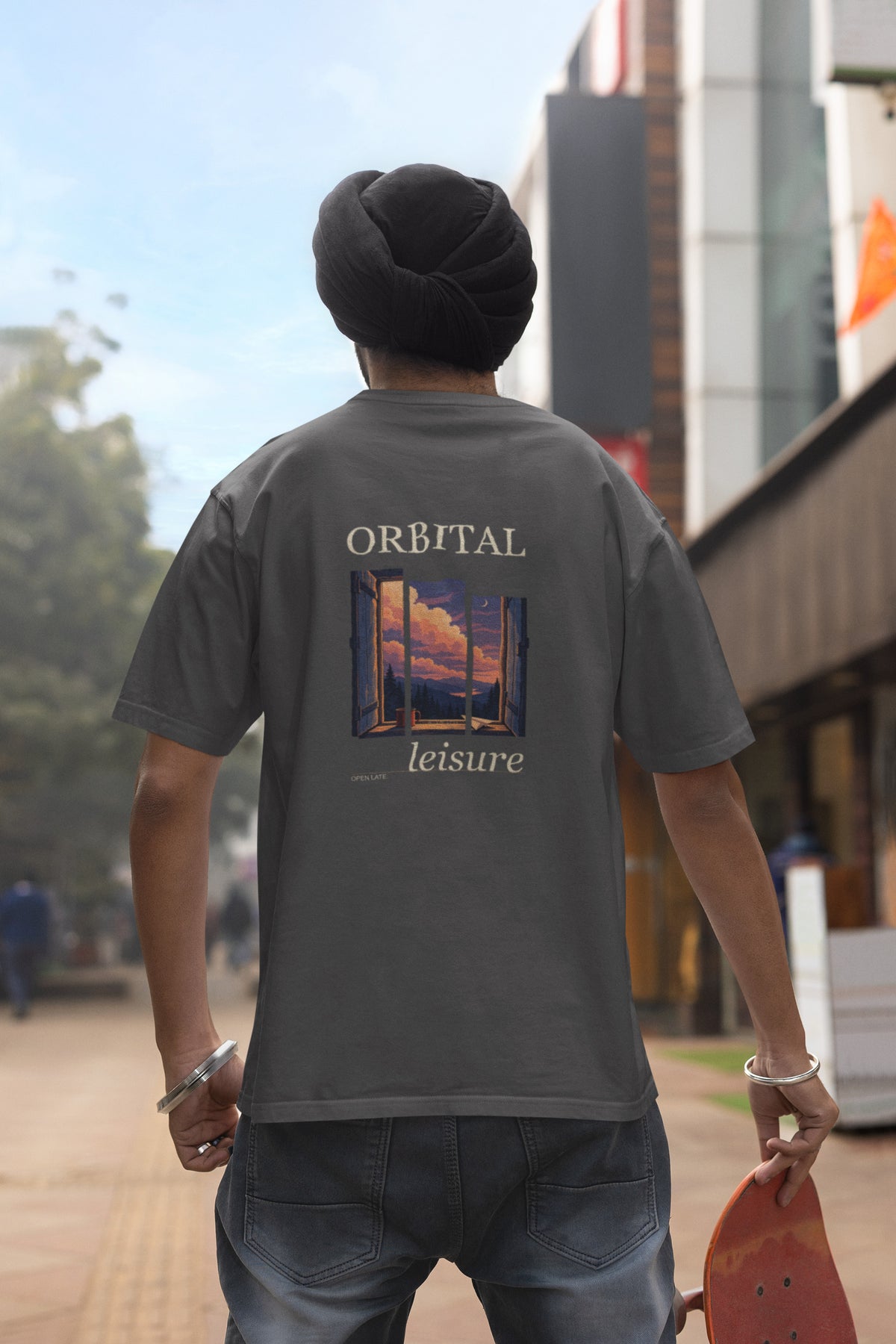 Open Late Garment-Dyed Tee in Graphite, worn by a man seen from behind in a turban on a busy street, showing the large print on the back. Digital product mockup.