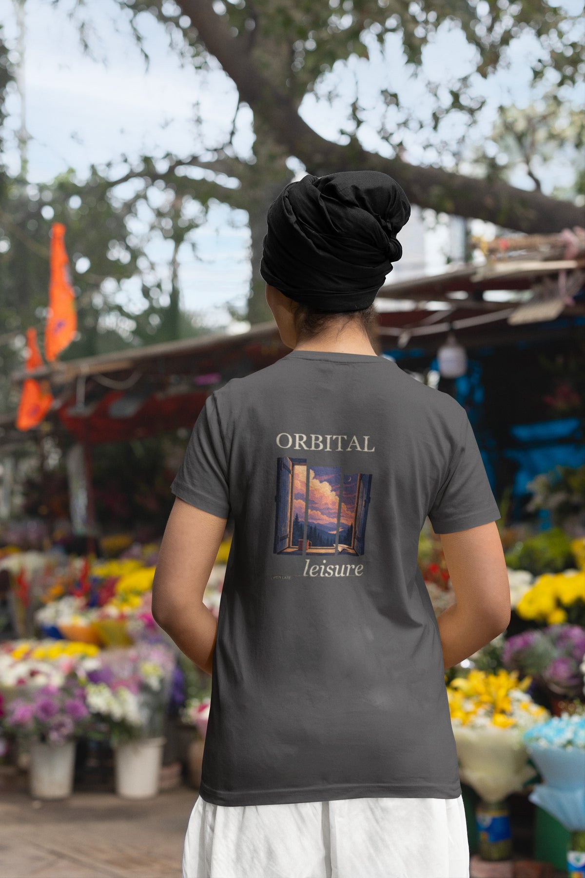 Open Late Garment-Dyed Tee in Graphite, worn by a woman seen from behind in a turban by a flower market, showing the large print on the back. Digital product mockup.