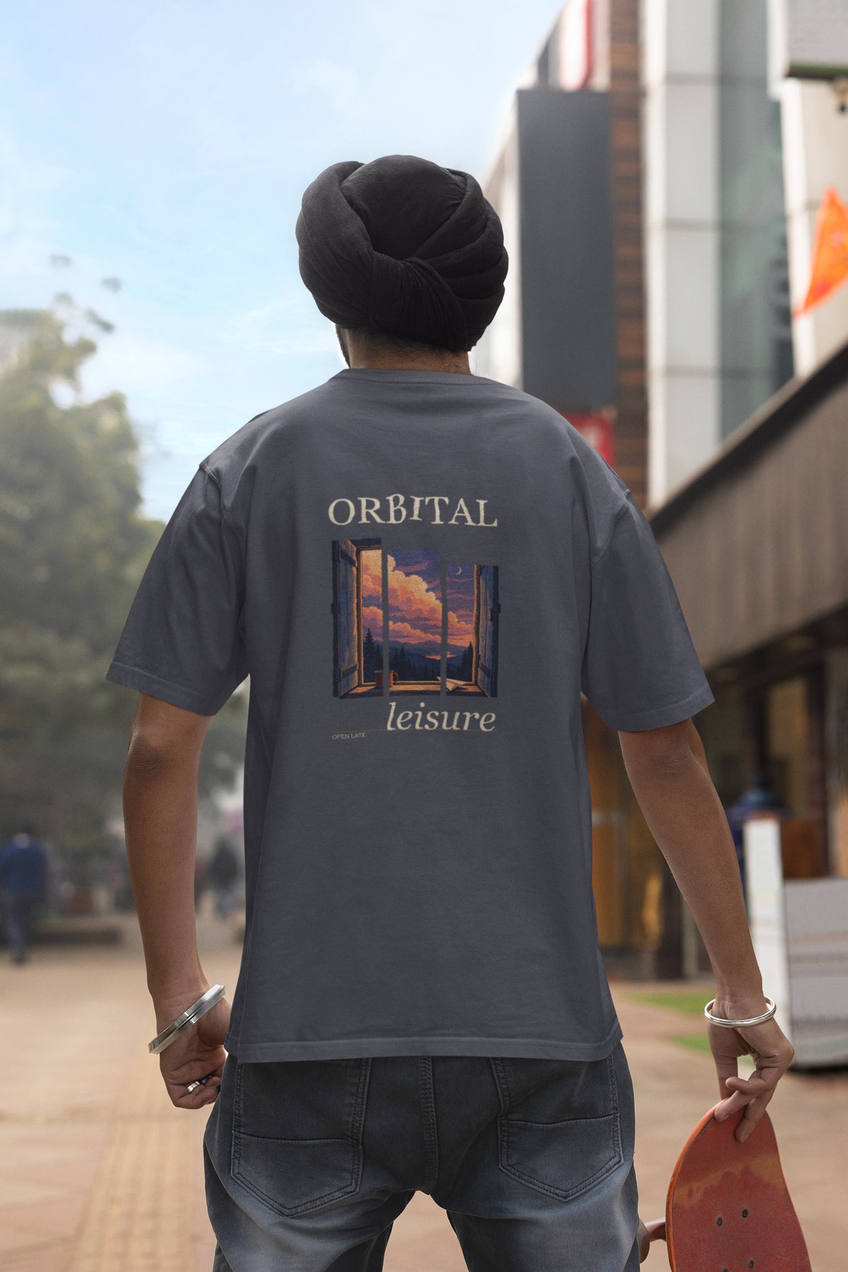Open Late Garment-Dyed Tee in Navy, worn by a man seen from behind in a turban on a busy street, showing the large print on the back. Digital product mockup.