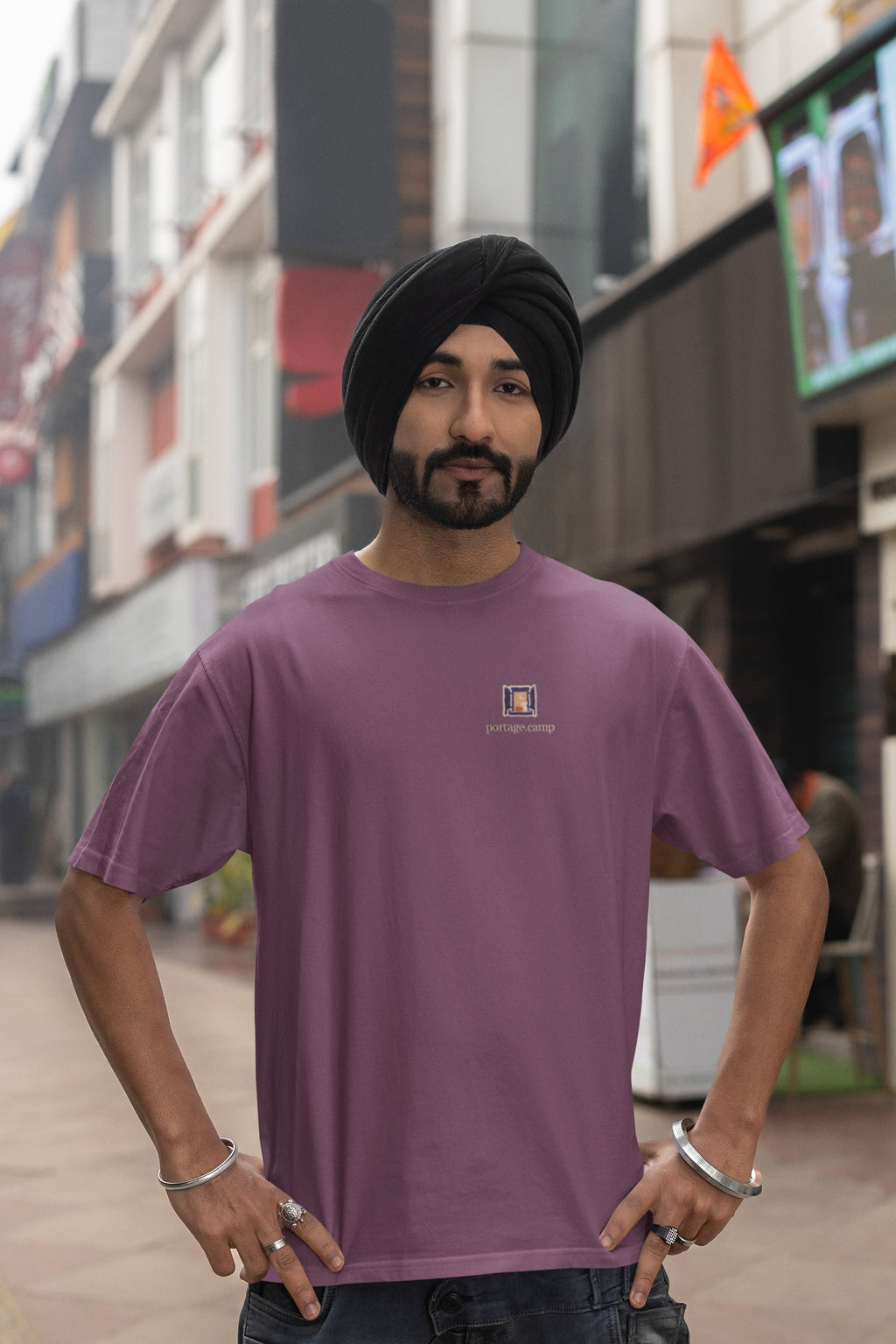 Open Late Garment-Dyed Tee in Berry, worn by a man in a turban on a busy street, with the Open Late logo and portage.camp on the chest. Digital product mockup.