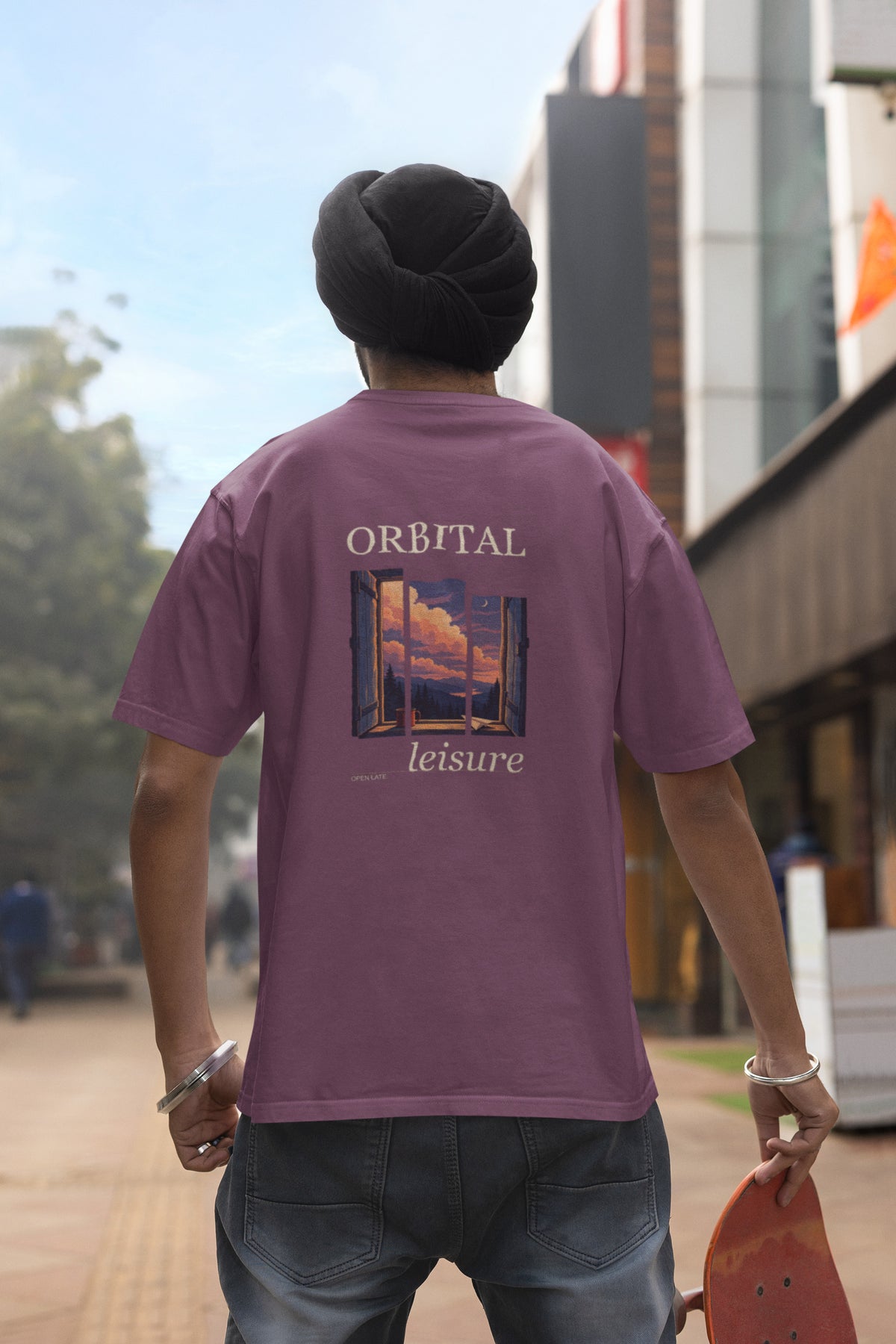 Open Late Garment-Dyed Tee in Berry, worn by a man seen from behind in a turban on a busy street, showing the large print on the back. Digital product mockup.
