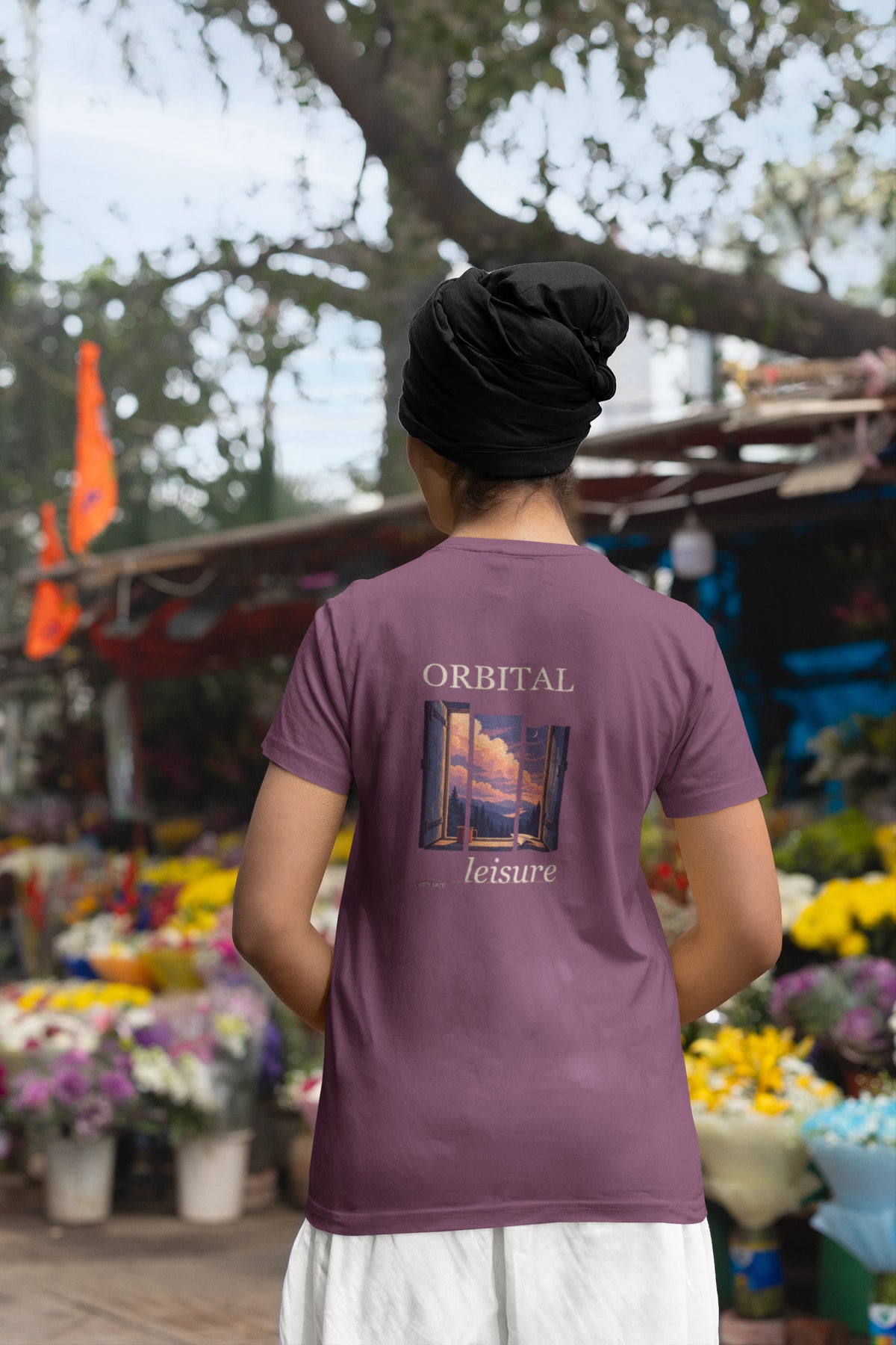 Open Late Garment-Dyed Tee in Berry, worn by a woman seen from behind in a turban by a flower market, showing the large print on the back. Digital product mockup.